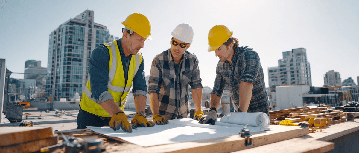 Three construction contractors in yellow hard hats reviewing architectural blueprints on rooftop with city skyline background - professional subcontractor team planning